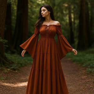 Puede incluir: Un vestido largo color &oacute;xido con hombros descubiertos y mangas largas acampanadas. El vestido presenta una falda escalonada y un detalle de lazo en el escote. La foto est&aacute; tomada al aire libre en un bosque.