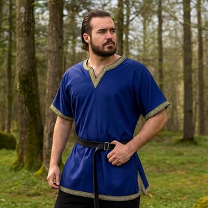 K&ouml;nnte beinhalten: Ein blaues Tunika mit goldenen Verzierungen am Ausschnitt und an den &Auml;rmeln sowie einem schwarzen G&uuml;rtel. Die Tunika hat einen V-Ausschnitt und kurze &Auml;rmel. Die Person steht in einem Wald.