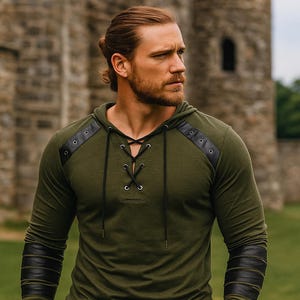 May include: An olive green hooded shirt with black leather accents on the shoulders and sleeves. The shirt features a lace-up neckline and is made of a soft, comfortable material. The model is standing in front of a stone building.