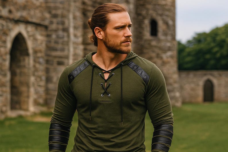 May include: An olive green hooded shirt with black leather accents on the shoulders and sleeves. The shirt features a lace-up neckline and is made of a soft, comfortable material. The model is standing in front of a stone building.