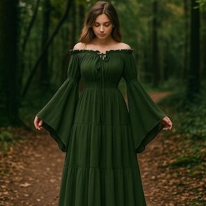 Puede incluir: Un vestido largo verde oscuro con hombros descubiertos, falda escalonada y mangas acampanadas. El vestido tiene un cuello con cord&oacute;n y se usa en un entorno forestal con un camino en el fondo. El vestido es de color s&oacute;lido.