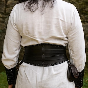 以下が含まれることがあります： 黒い革のコルセットベルト。編み込みのエッジと垂直のストラップが付いており、白い長袖シャツの上に着用されています。ベルトの側面には小さなポーチが付いています。袖には黒い袖口が付いています。