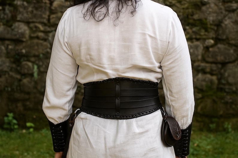 以下が含まれることがあります： 黒い革のコルセットベルト。編み込みのエッジと垂直のストラップが付いており、白い長袖シャツの上に着用されています。ベルトの側面には小さなポーチが付いています。袖には黒い袖口が付いています。