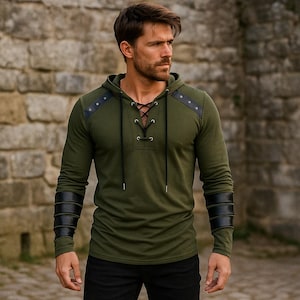 May include: Olive green hooded long-sleeve shirt with black leather accents on the shoulders and forearms. The shirt features a lace-up neckline and is paired with black pants. The model is standing in front of a stone wall.
