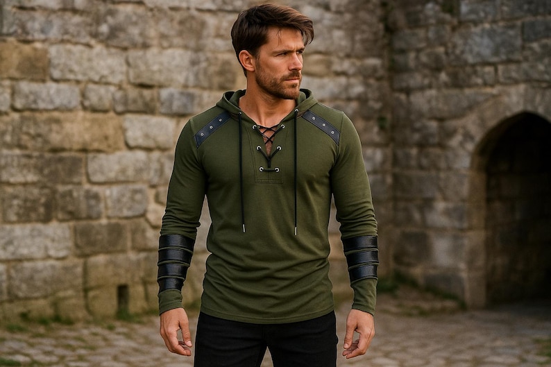 May include: Olive green hooded long-sleeve shirt with black leather accents on the shoulders and forearms. The shirt features a lace-up neckline and is paired with black pants. The model is standing in front of a stone wall.