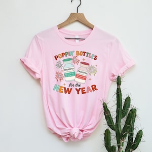 May include: A pale pink t-shirt with the words "POPPIN' BOTTLES for the NEW YEAR" in various colours. The design includes illustrations of two medicine bottles labelled "PROPOFOL" with fireworks and star accents.