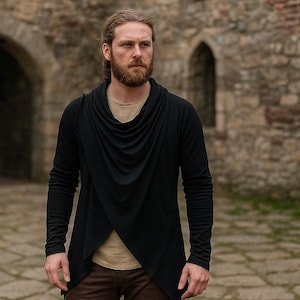 Peut inclure: Haut noir à manches longues avec un col drapé et un ourlet asymétrique. Le haut est superposé à une chemise beige et associé à un pantalon marron. Le modèle se tient devant un bâtiment en pierre.