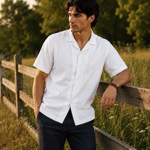 May include: A short-sleeved white button-down shirt with a camp collar and a straight hem, paired with dark navy trousers. The shirt features a visible button placket down the center and a textured fabric finish, worn with a simple brown wristband.