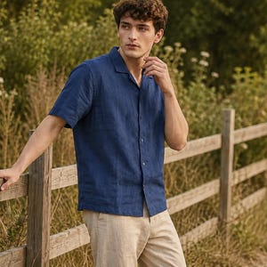 May include: A short-sleeved navy blue button-down shirt with a notched collar and a straight hem, paired with light beige trousers. The shirt features a visible texture and a central row of matching buttons, worn untucked over the pants.