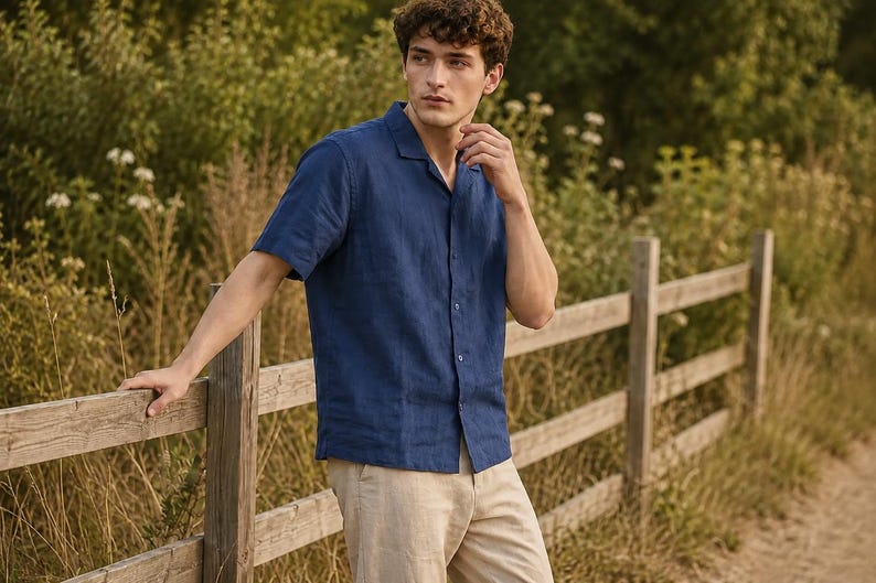 May include: A short-sleeved navy blue button-down shirt with a notched collar and a straight hem, paired with light beige trousers. The shirt features a visible texture and a central row of matching buttons, worn untucked over the pants.
