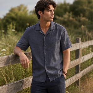 May include: A short-sleeved button-down shirt in a dark navy blue color, featuring a classic open camp collar, a straight hemline, and a textured fabric finish, worn with dark navy blue trousers and a brown leather wristband accessory.