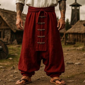 May include: Burgundy-colored, loose-fitting pants with a high waist and decorative buttons. The pants are part of a traditional outfit, paired with a white shirt. The image is set in a rustic, outdoor environment.