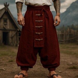 May include: Burgundy-colored, wide-legged pants with a high waist and decorative clasps. The pants are part of a historical or fantasy-themed outfit. The person is wearing sandals. The background features a rustic village setting with wooden buildings and mountains.