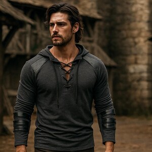 May include: A dark gray hooded shirt with leather accents on the shoulders and sleeves. The shirt features a lace-up neckline and is worn by a person in an outdoor setting. The sleeves have black leather bands.