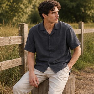 May include: A dark charcoal gray short-sleeved button-down shirt with a notched collar and a textured fabric finish, paired with light beige trousers. The shirt features a straight hem and a button-front closure, worn untucked over the pants.