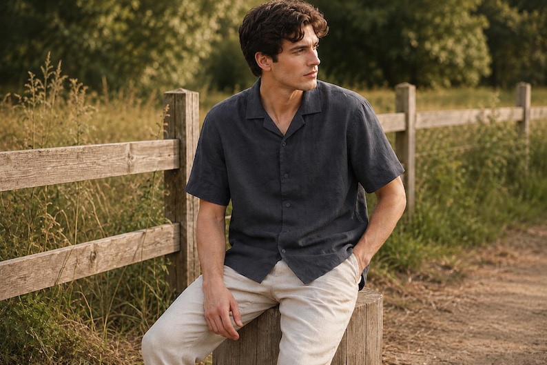 May include: A dark charcoal gray short-sleeved button-down shirt with a notched collar and a textured fabric finish, paired with light beige trousers. The shirt features a straight hem and a button-front closure, worn untucked over the pants.