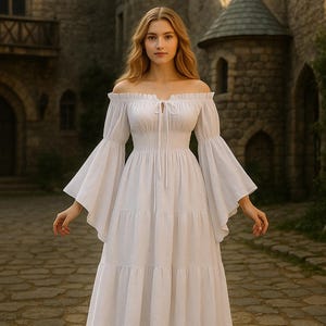 Puede incluir: Un vestido blanco con hombros descubiertos y mangas largas acampanadas. El vestido presenta una cintura ce&ntilde;ida y una falda escalonada. La modelo est&aacute; de pie en un camino de piedra frente a un castillo.