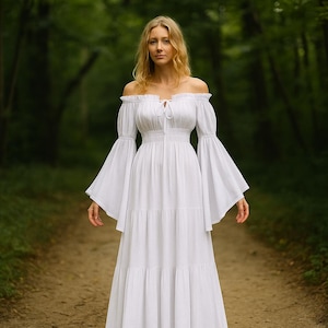 Puede incluir: Un vestido blanco con hombros descubiertos y mangas largas acampanadas. El vestido tiene un corpi&ntilde;o ajustado y una falda escalonada. La mujer est&aacute; de pie en un camino en un bosque. El vestido es de tela ligera.