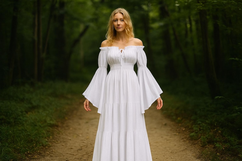 Puede incluir: Un vestido blanco con hombros descubiertos y mangas largas acampanadas. El vestido tiene un corpi&ntilde;o ajustado y una falda escalonada. La mujer est&aacute; de pie en un camino en un bosque. El vestido es de tela ligera.