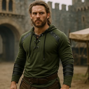 May include: An olive green hooded shirt with black leather accents on the shoulders and sleeves. The shirt features a lace-up neckline and is paired with a brown belt and pants. The background shows a stone building and a tent.