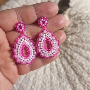 Könnte beinhalten: Ein Paar tropfenförmige Ohrringe mit aufwendigen Perlenarbeiten in den Farben Pink, Hellrosa und Weiß. Jeder Ohrring besteht aus einem kleinen runden Stecker, der mit einem größeren, tropfenförmigen Anhänger mit gewelltem Rand verbunden ist.