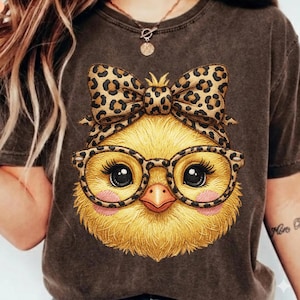 May include: A brown t-shirt featuring a cartoon chick with a leopard print bow, glasses, and headband. The chick has yellow feathers, pink cheeks, and large black eyes. The overall design is whimsical and playful.