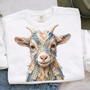 May include: White sweatshirt featuring a detailed illustration of a goat's head. The goat's face is adorned with patches of fabric in floral, plaid, and denim patterns. The goat has brown eyes, tan fur, and curved horns. The sweatshirt has a crew neck.