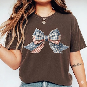 Op de afbeelding: Bruin t-shirt met een patriottisch strikdesign. De strik heeft blauw-wit ruitjes, rood-witte strepen en blauwe delen met witte sterren. Het shirt is een casual stijl.
