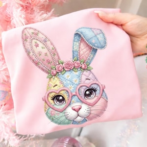 May include: A pink t-shirt featuring a colorful patchwork bunny design. The bunny has heart-shaped glasses, a floral crown, and a quilted pattern in pastel colors. The design is detailed and whimsical, perfect for Easter.