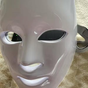 May include: A white, glossy mask with a smooth surface. The mask has eye openings and a mouth, with a strap on the side. The mask is designed to cover the entire face. The mask is likely for costume or theatrical use.