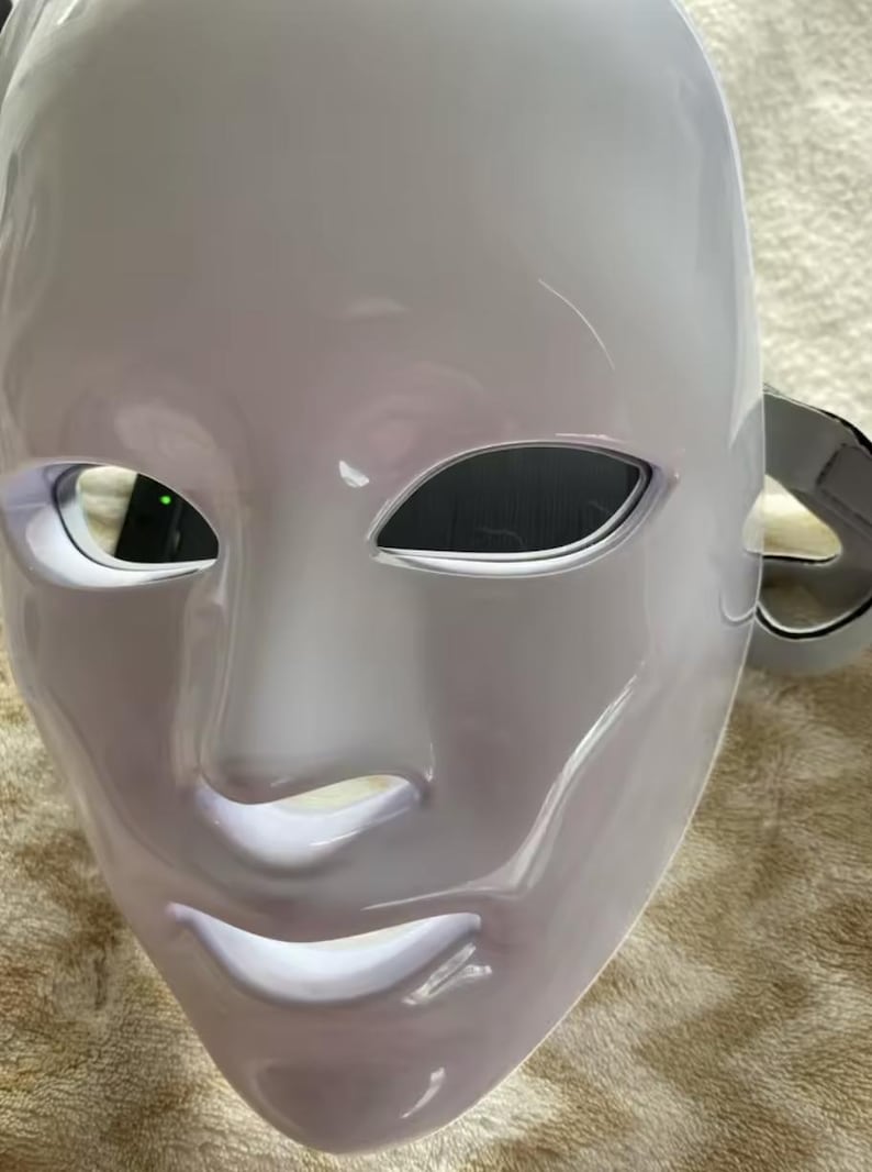 May include: A white, glossy mask with a smooth surface. The mask has eye openings and a mouth, with a strap on the side. The mask is designed to cover the entire face. The mask is likely for costume or theatrical use.