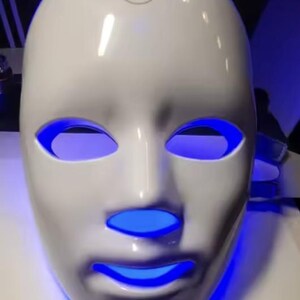 May include: A white LED light therapy mask with blue light emanating from the eye and mouth openings. The mask has a smooth, glossy finish and a small logo on the forehead area. The mask is designed to fit the contours of a face.
