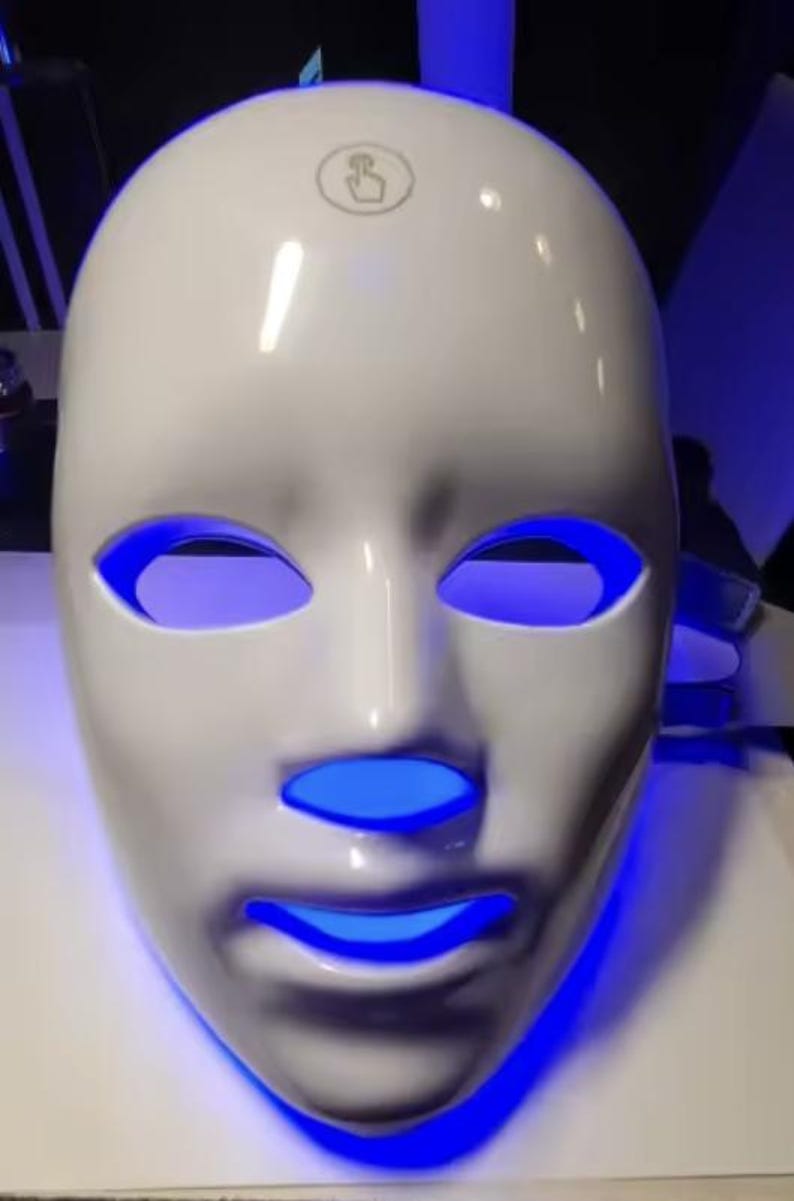May include: A white LED light therapy mask with blue light emanating from the eye and mouth openings. The mask has a smooth, glossy finish and a small logo on the forehead area. The mask is designed to fit the contours of a face.