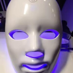 May include: A white, face-shaped LED mask with glowing purple light emanating from the eye and mouth openings. The mask has a smooth, glossy finish and a touch icon on the forehead.