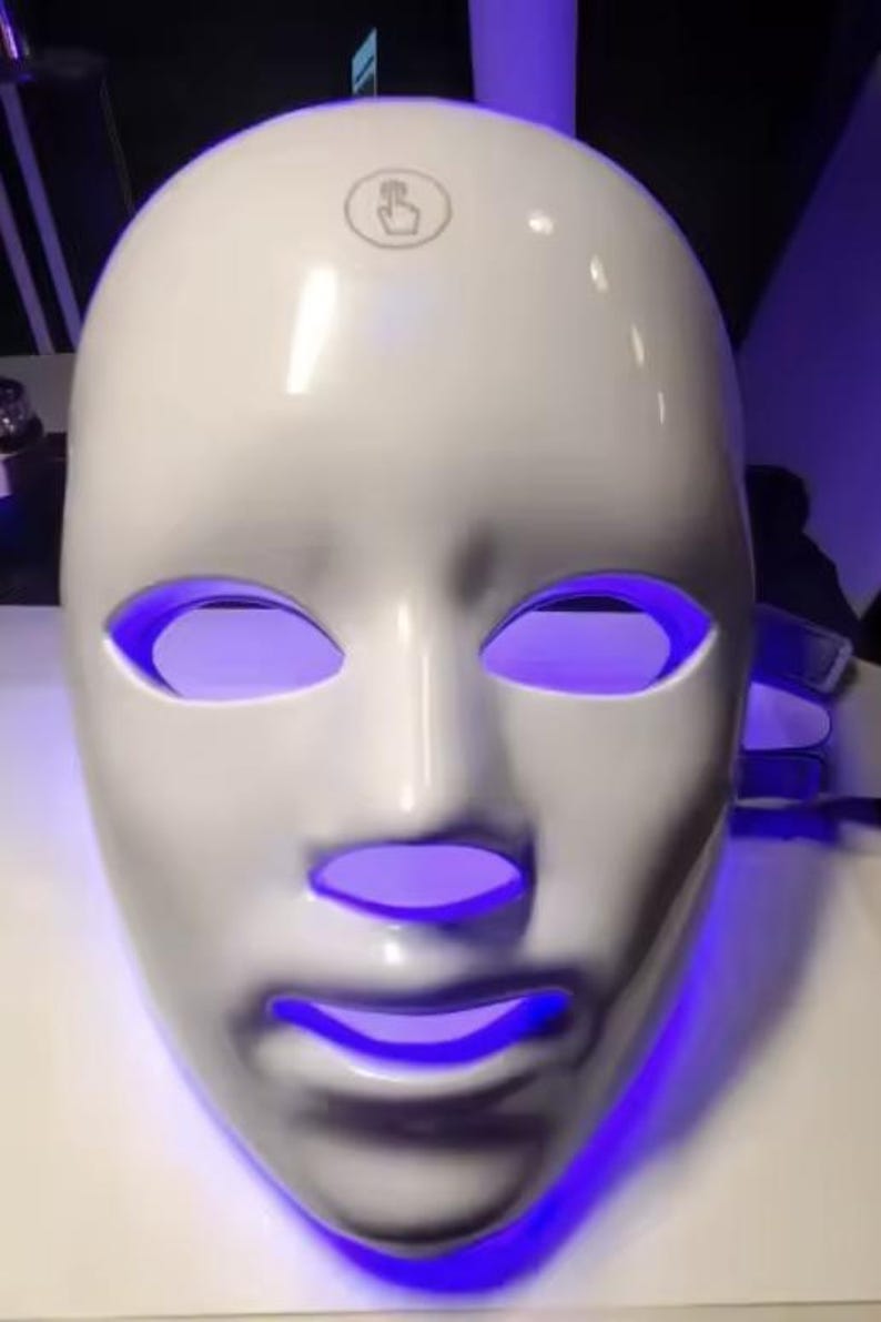 May include: A white, face-shaped LED mask with glowing purple light emanating from the eye and mouth openings. The mask has a smooth, glossy finish and a touch icon on the forehead.