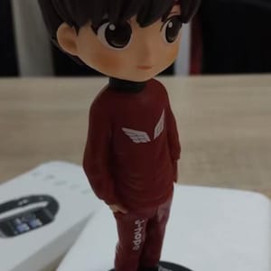 May include: A figurine of a person with dark hair and a red outfit. The figure is standing on a black base. The red outfit has white wing designs on the chest and the text "j-hope" on the leg.