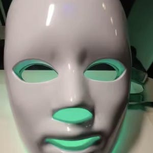 May include: A white LED face mask with eye, nose, and mouth openings. The mask has a smooth surface and a touch icon on top. The openings glow with a green light, suggesting a skincare or beauty product.