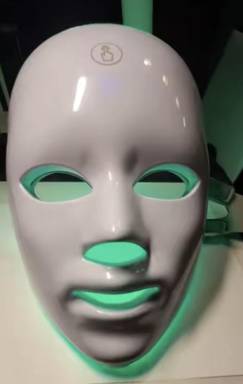 May include: A white LED face mask with eye, nose, and mouth openings. The mask has a smooth surface and a touch icon on top. The openings glow with a green light, suggesting a skincare or beauty product.