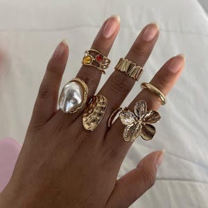 Peut inclure: Une main ornée de plusieurs bagues dorées. Les bagues présentent divers motifs, notamment une grosse perle, des bandes texturées, une bague florale et des bagues avec des pierres colorées. Les bagues varient en taille et en style.