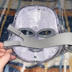 May include: A clear, LED light therapy mask with gray adjustable straps. The mask has a contoured design with eye openings and is filled with numerous small lights. The mask is designed to fit the face and is likely used for skincare treatments.