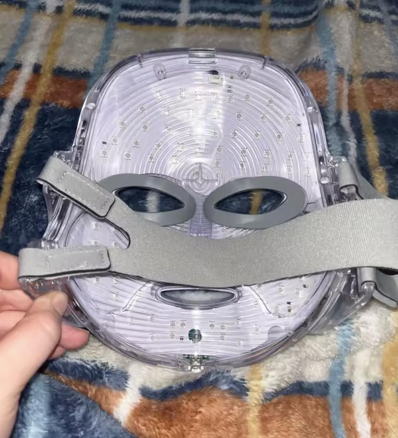 May include: A clear, LED light therapy mask with gray adjustable straps. The mask has a contoured design with eye openings and is filled with numerous small lights. The mask is designed to fit the face and is likely used for skincare treatments.