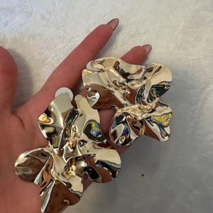 Peut inclure: Une paire de grandes boucles d'oreilles dorées avec un design abstrait et organique. Les boucles d'oreilles ont une surface réfléchissante et texturée, et un fermoir à tige. Les boucles d'oreilles sont présentées sur un fond clair.