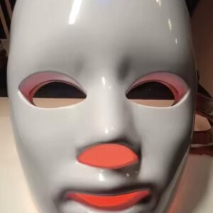 May include: A white LED light therapy mask with red light visible around the eyes, nose, and mouth. The mask has a smooth, glossy finish and a touch icon on the forehead.