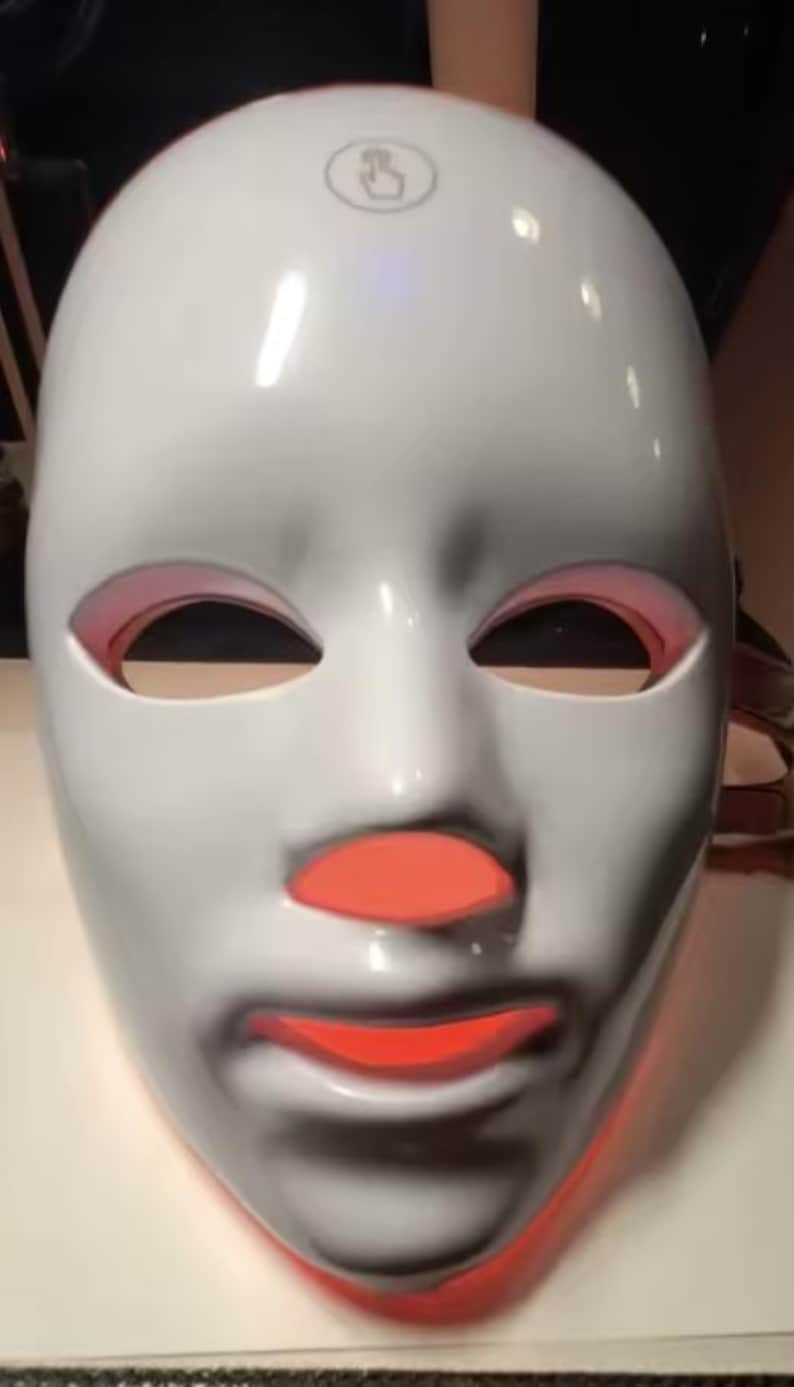 May include: A white LED light therapy mask with red light visible around the eyes, nose, and mouth. The mask has a smooth, glossy finish and a touch icon on the forehead.