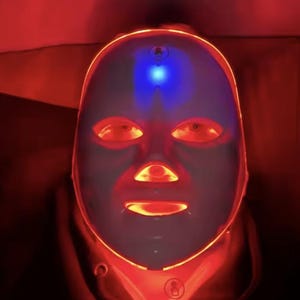 May include: A futuristic-looking LED light therapy mask. The mask is white with red and blue lights illuminating the eye, nose, and mouth areas. The mask is designed to cover the entire face and is surrounded by a red glow.