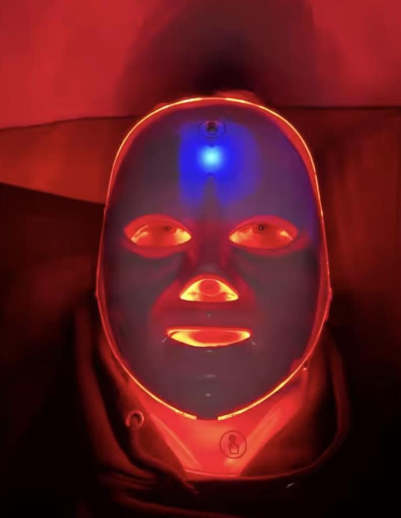 May include: A futuristic-looking LED light therapy mask. The mask is white with red and blue lights illuminating the eye, nose, and mouth areas. The mask is designed to cover the entire face and is surrounded by a red glow.