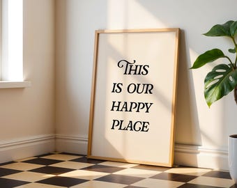 May include: A framed print with the words "This is our Happy Place" in black script. The frame is light wood, and the background is a cream color. The print is leaning against a wall next to a potted plant.