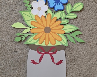 May include: A papercraft bouquet of flowers in a mason jar shape. The bouquet features blue, white, and orange flowers with green leaves. A red ribbon is tied around the jar. The background is a neutral carpet.
