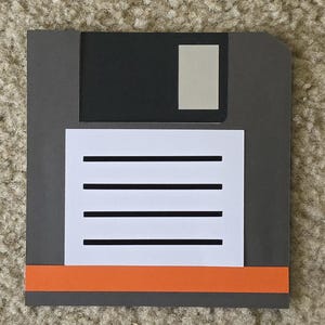 May include: A retro-style artwork depicting a floppy disk. The design features a gray rectangular base with a black top section and a white label area. The label has four black horizontal lines, and an orange stripe at the bottom.