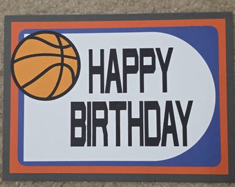 May include: A rectangular birthday card with a basketball graphic. The card features a basketball on the left, with the words "HAPPY BIRTHDAY" in bold black letters. The card has a gray border, orange and blue accents.